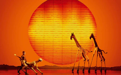 Stage performance of The Lion King with actors as animals against a sunset backdrop.