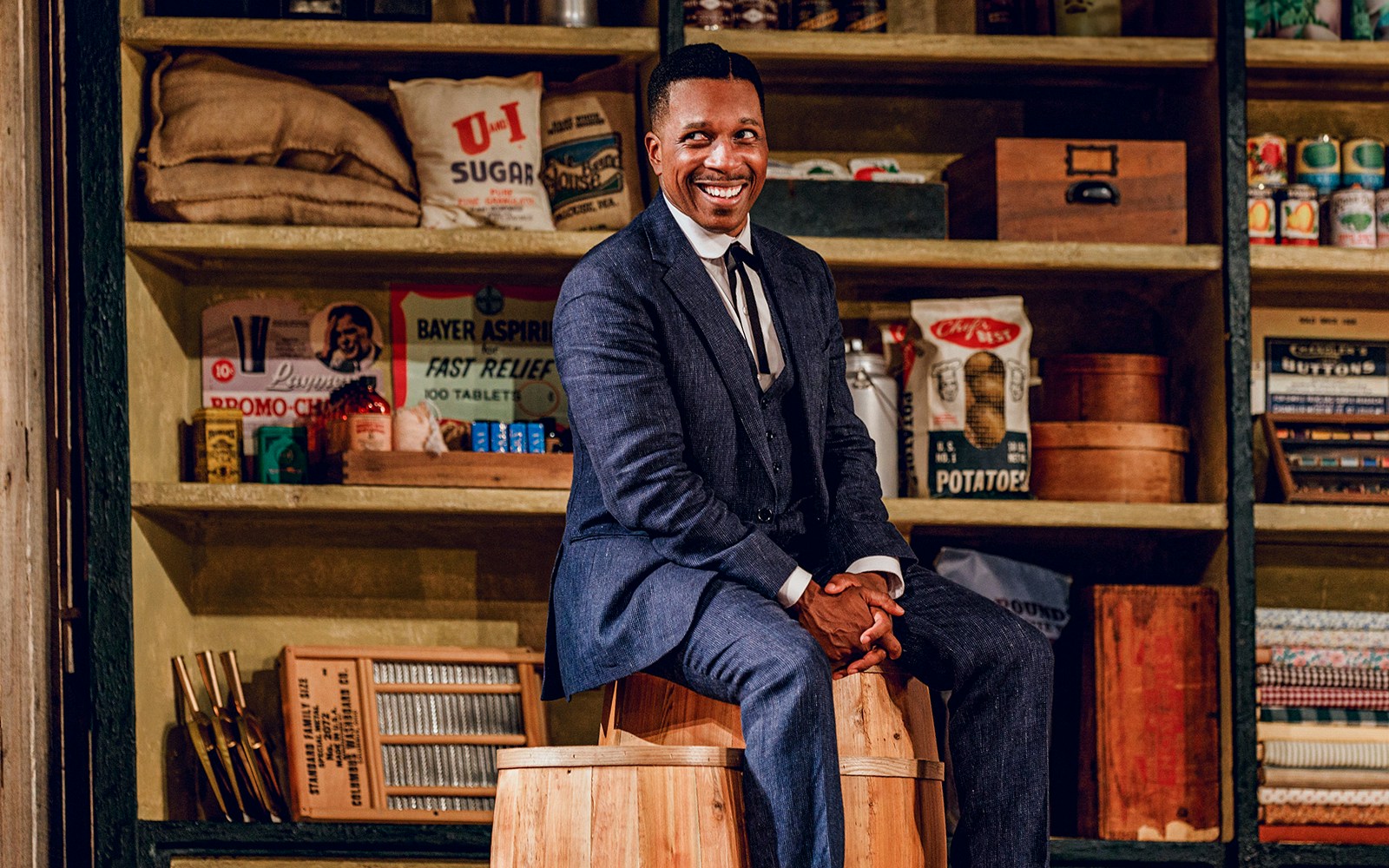 Man in a suit sitting on a wooden barrel in a vintage store setting, related to "Purlie Victorious.