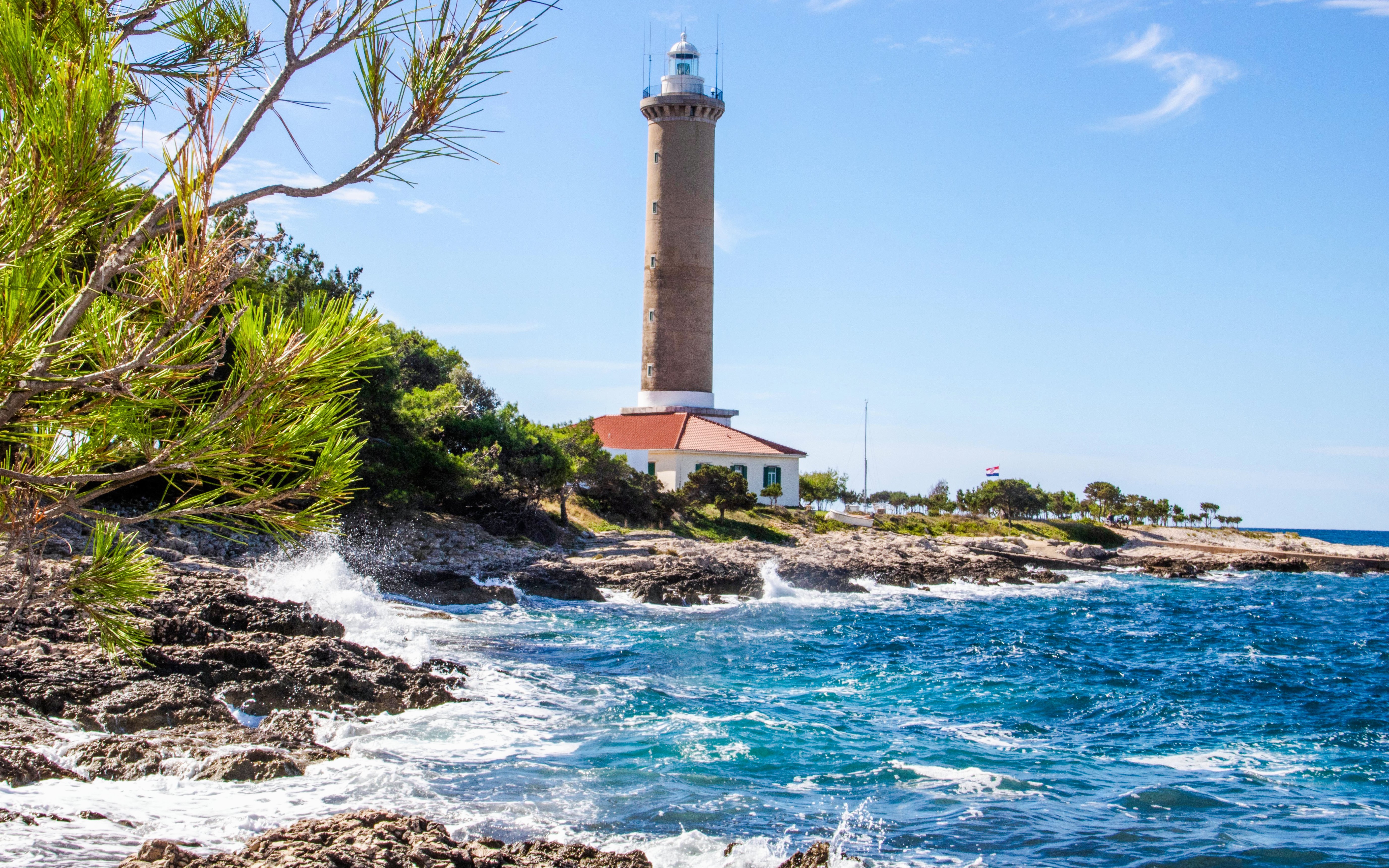 Veli Rat Lighthouse on rocky Adriatic coast, Croatia, with surrounding greenery.