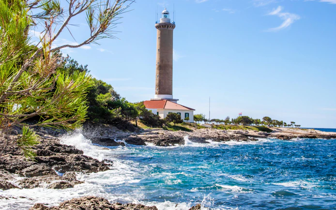 Veli Rat Lighthouse on rocky Adriatic coast, Croatia, with surrounding greenery.