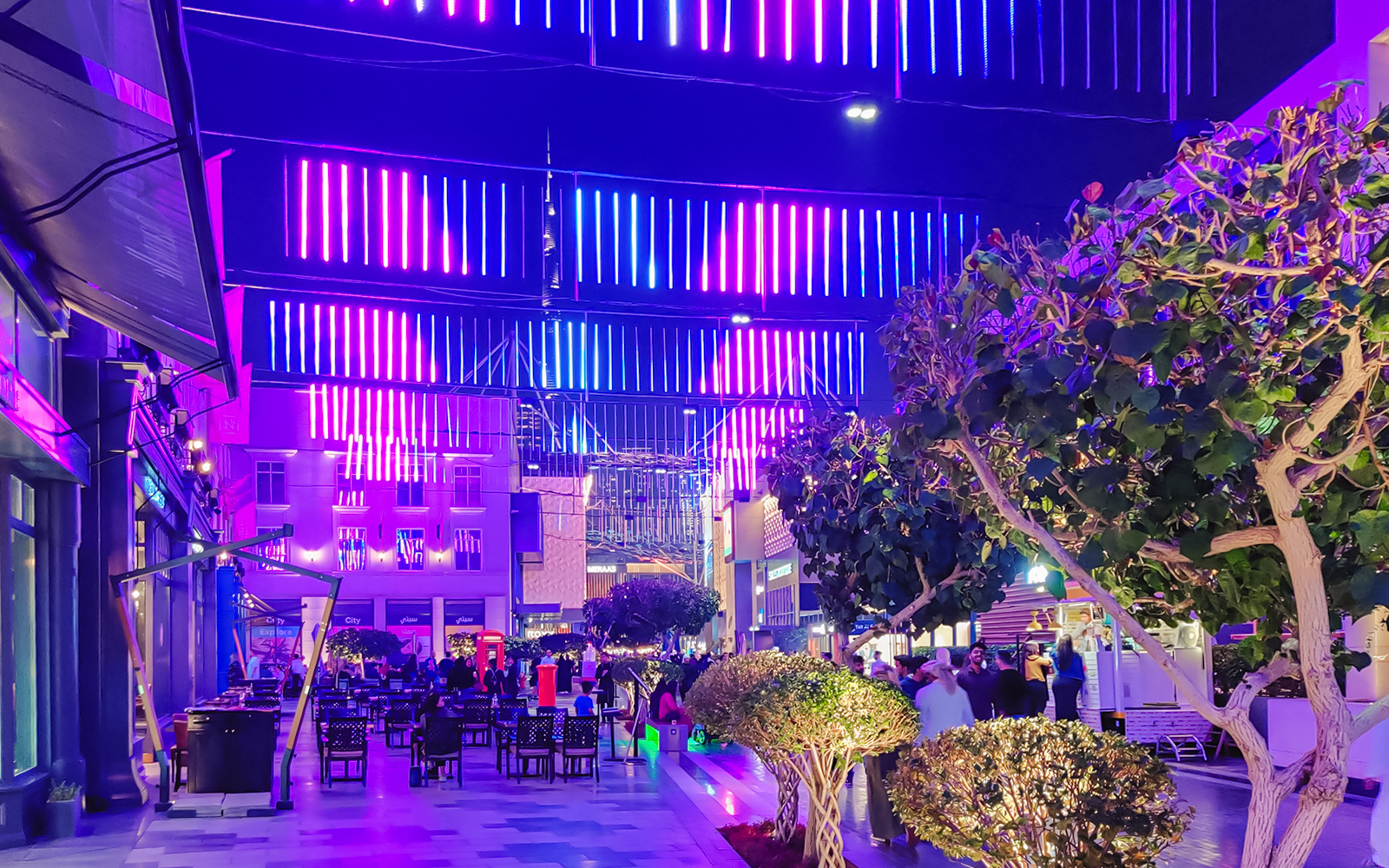 Neon-lit street with shops and outdoor seating, vibrant night scene.