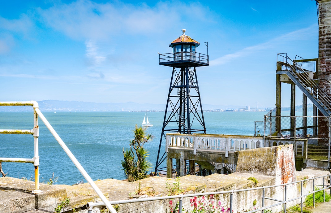 A scenic view of Alcatraz Island, showing the historic guard tower, a white railing, and steps leading down towards the blue waters of San Francisco Bay