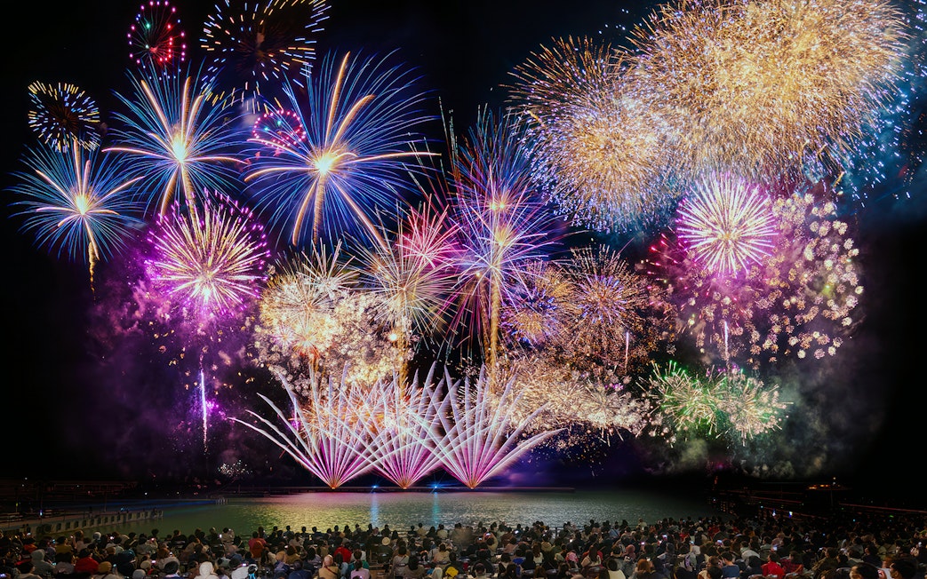 Fireworks display over water at Huis Ten Bosch, Japan, viewed by a large crowd.
