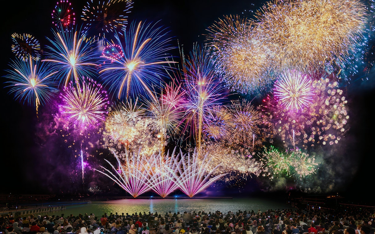 Fireworks display over water at Huis Ten Bosch, Japan, viewed by a large crowd.