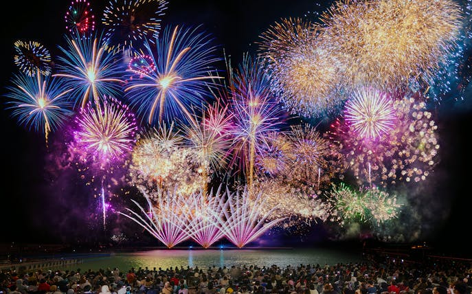 Fireworks display over water at Huis Ten Bosch, Japan, viewed by a large crowd.