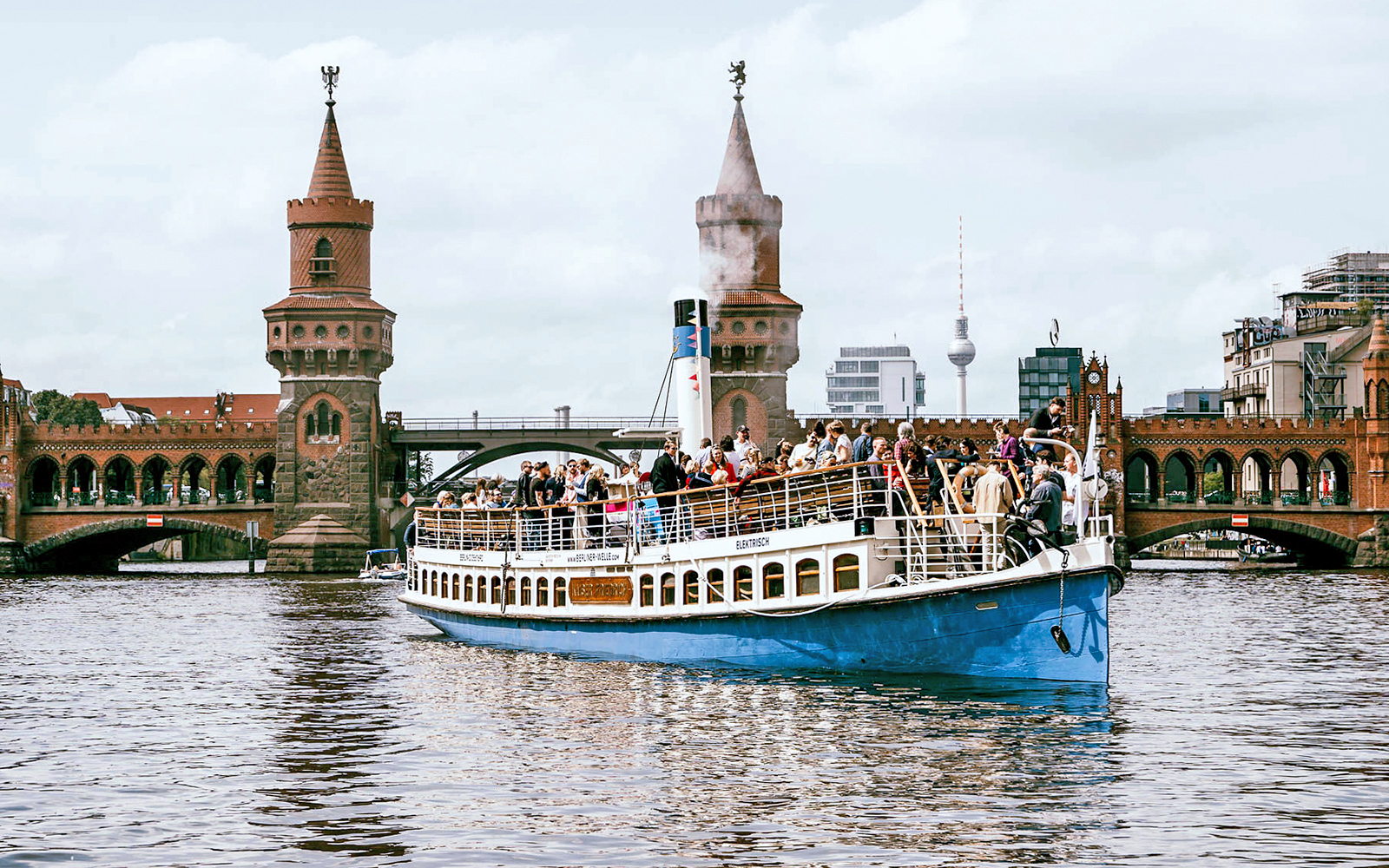 Tourists on a boat cruise past Berlin's Oberbaum Bridge with TV Tower in the background.