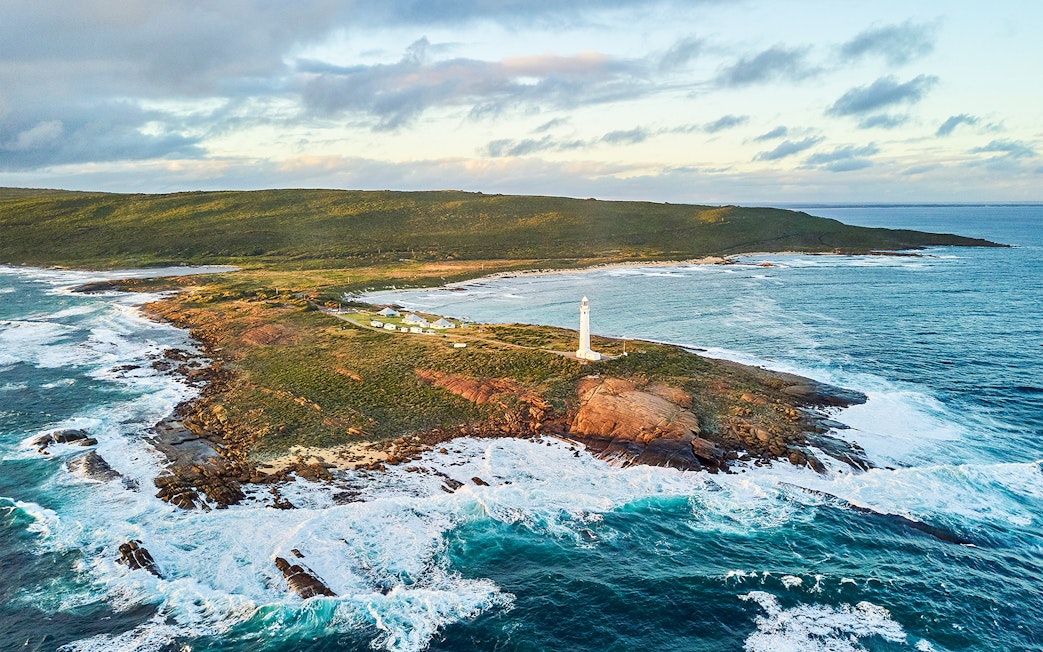 Aerial view of Cape Leeuwin Lighthouse on rocky coastal headland, surrounded by ocean waves.