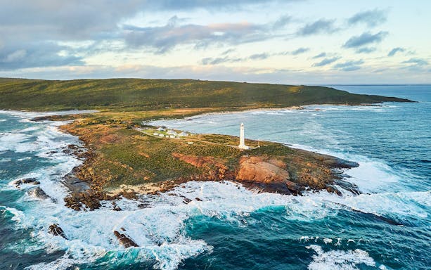 Aerial view of Cape Leeuwin Lighthouse on rocky coastal headland, surrounded by ocean waves.