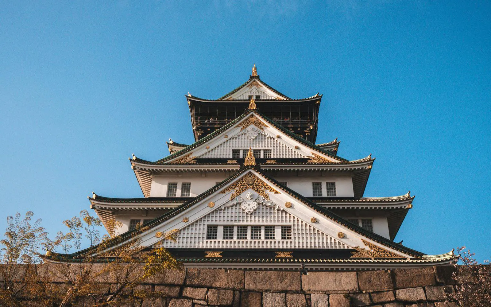 Osaka Castle with ornate architecture against a clear blue sky, surrounded by cherry blossoms.