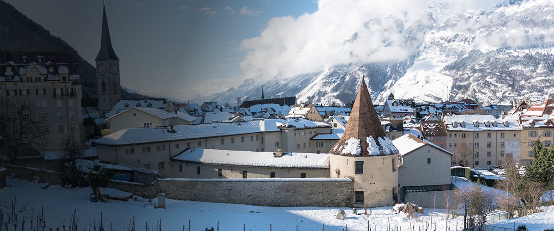 Snow-covered rooftops and church tower in Chur, Switzerland, with mountains in the background.