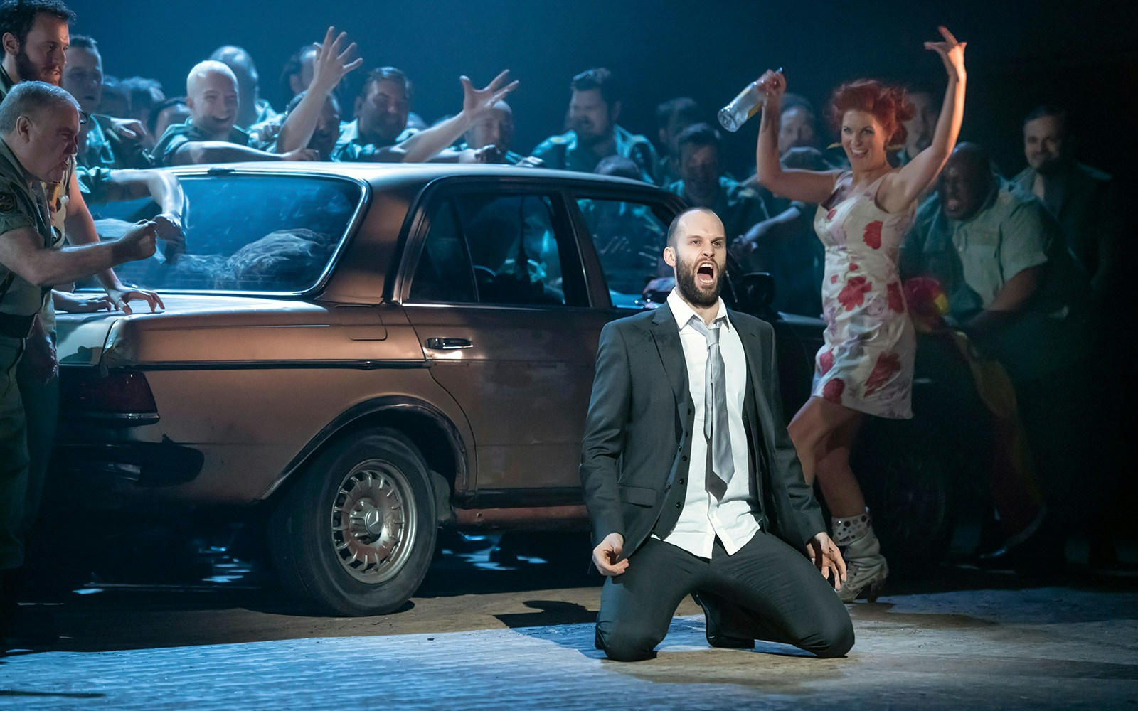 Opera performance of Carmen with a man kneeling and a woman dancing near a car.