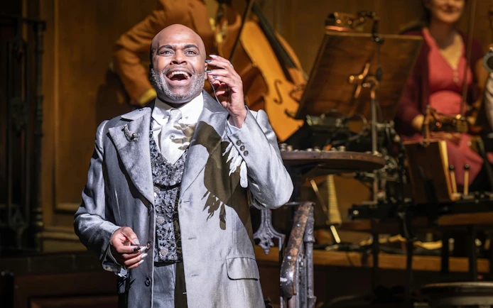 Performer on stage during Hadestown musical, West End, with musicians in the background.