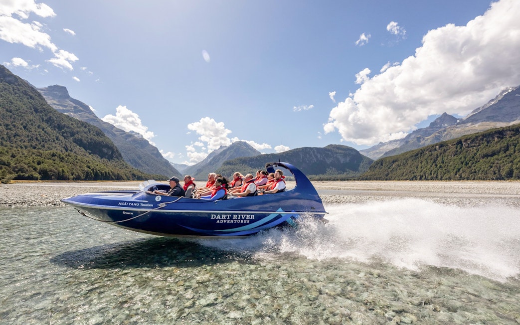 Jet boat on Dart River with passengers, surrounded by mountains and clear skies, New Zealand.