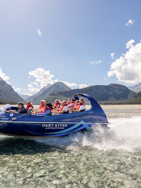 Jet boat on Dart River with passengers, surrounded by mountains and clear skies, New Zealand.