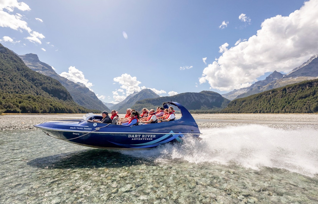 Jet boat on Dart River with passengers, surrounded by mountains and clear skies, New Zealand.