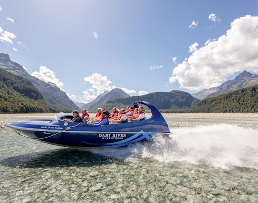 Jet boat on Dart River with passengers, surrounded by mountains and clear skies, New Zealand.