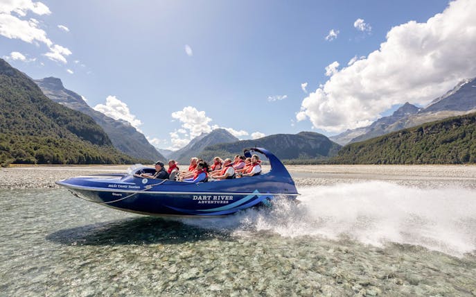 Jet boat on Dart River with passengers, surrounded by mountains and clear skies, New Zealand.