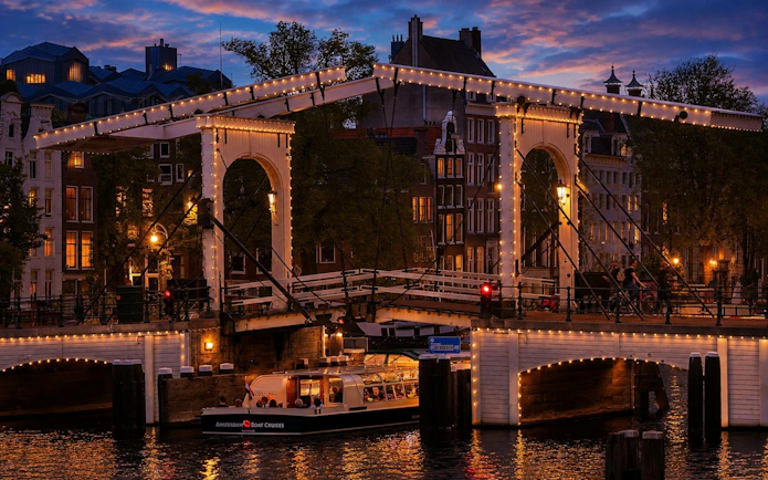 Amsterdam canal cruise boat under illuminated bridge at sunset.