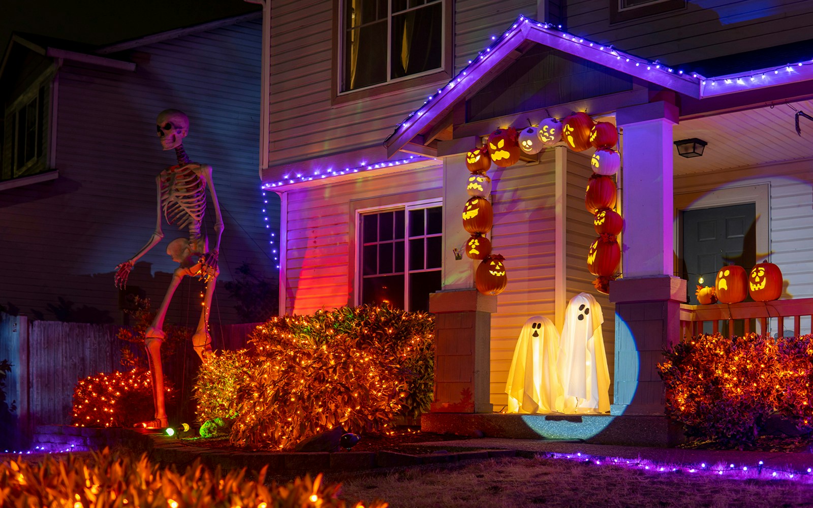 Halloween house with orange and purple lights, skeleton, pumpkins, and ghost decorations on porch.
