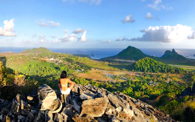 Person sitting on rocky viewpoint overlooking lush landscape and ocean, Noronha Piquinho Trail, Sao Paulo.