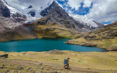Hiker walking by a turquoise lake with snow-capped mountains in Ausangate, Peru.
