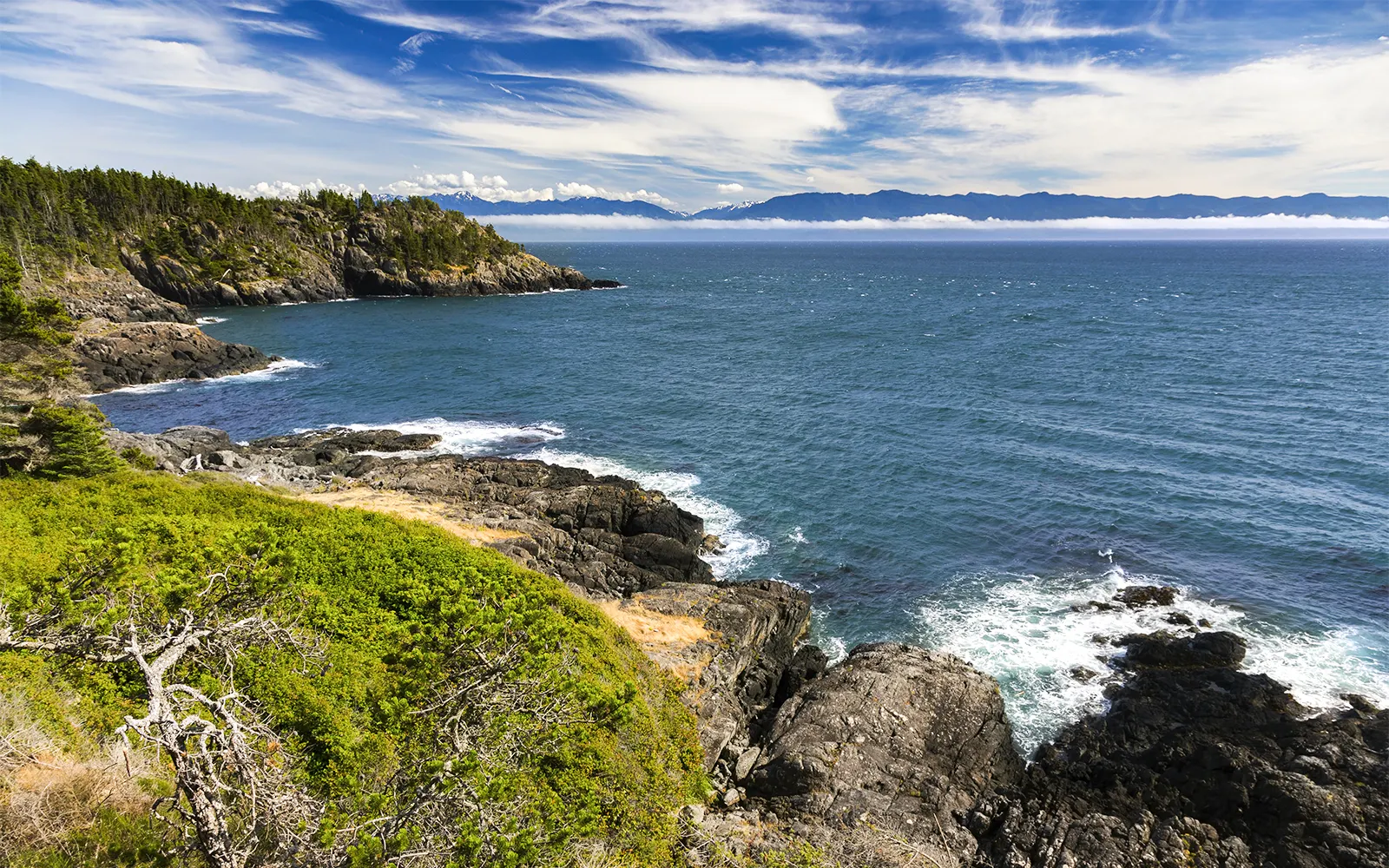 Sweeping view of Juan De Fuca Strait with rocky coastline and Olympic Peninsula mountains in the distance.