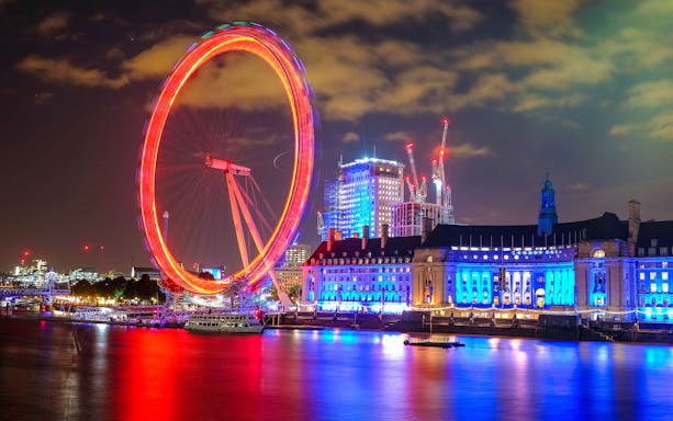 London Eye illuminated at night with city skyline and reflections on the Thames.