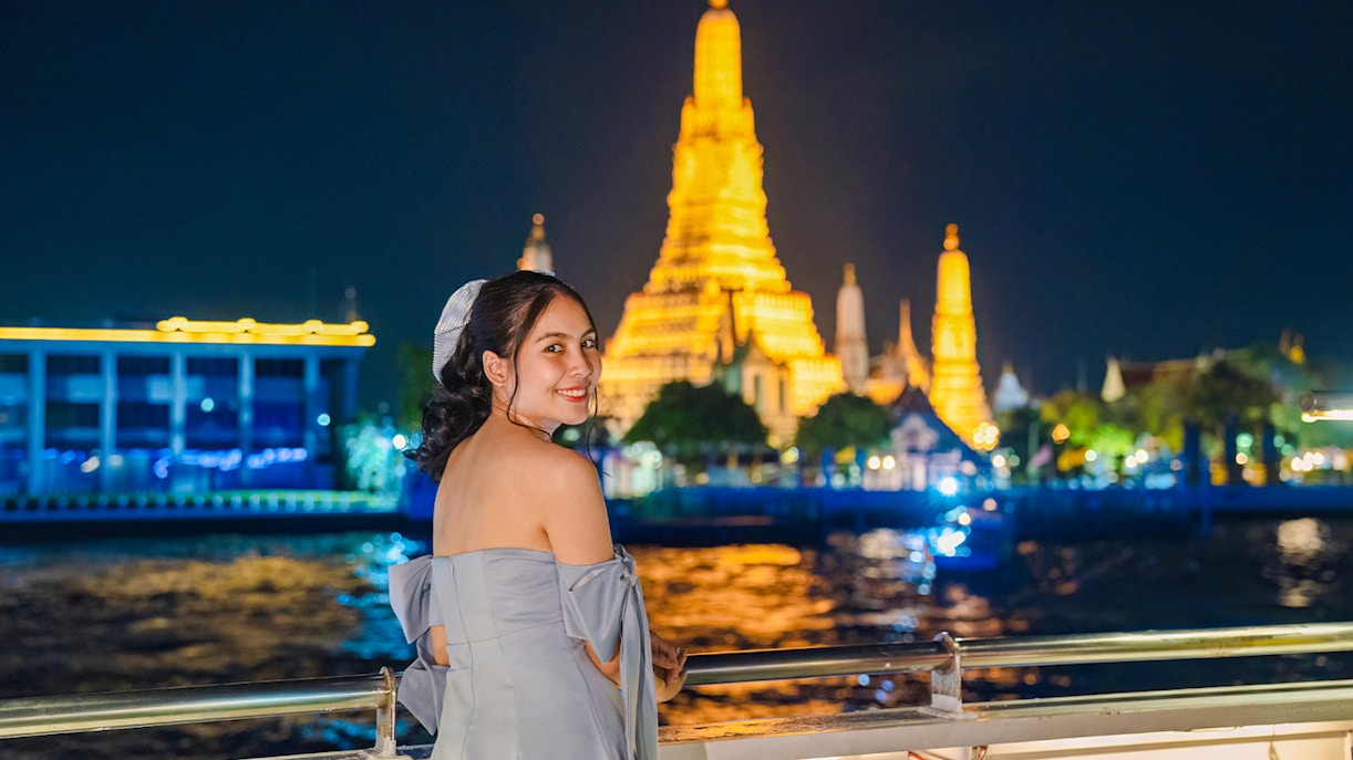 Woman enjoying a view of Wat Arun illuminated at night from a dinner cruise in Bangkok.
