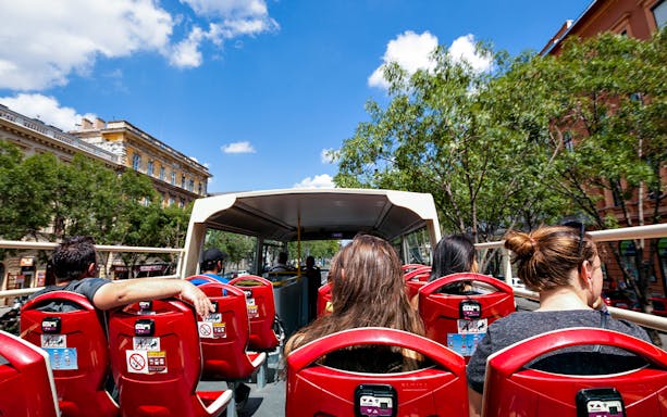 Open-top bus tour with passengers in a city setting, surrounded by trees and historic buildings.