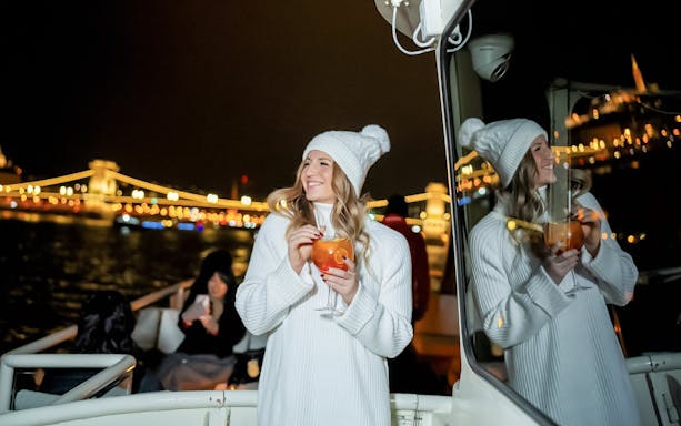 Guests enjoying drinks on a Budapest Danube River nighttime sightseeing cruise with city lights in the background.