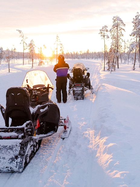 Snowmobiles on snowy trail at sunset in Lapland forest.