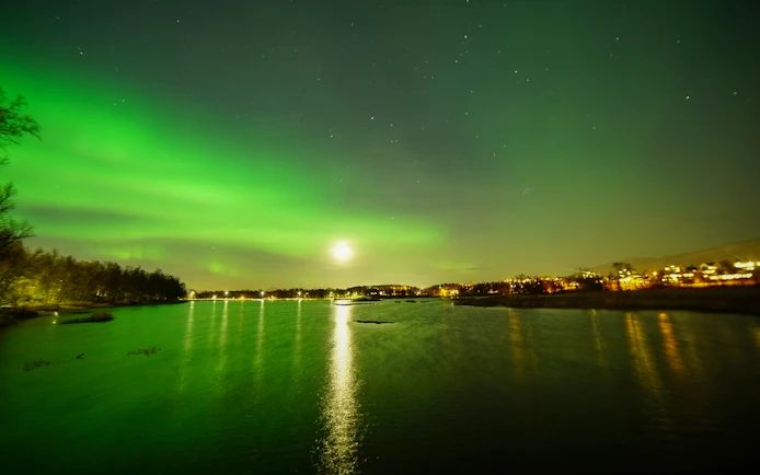 Aurora borealis over a calm lake with distant city lights reflecting on the water.