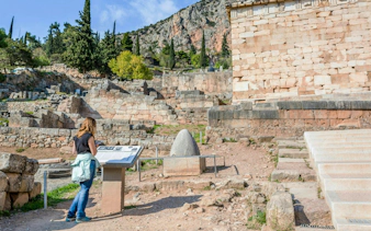 Visitor reading plaque at Delphi ruins, Greece, with ancient stone structures in background.