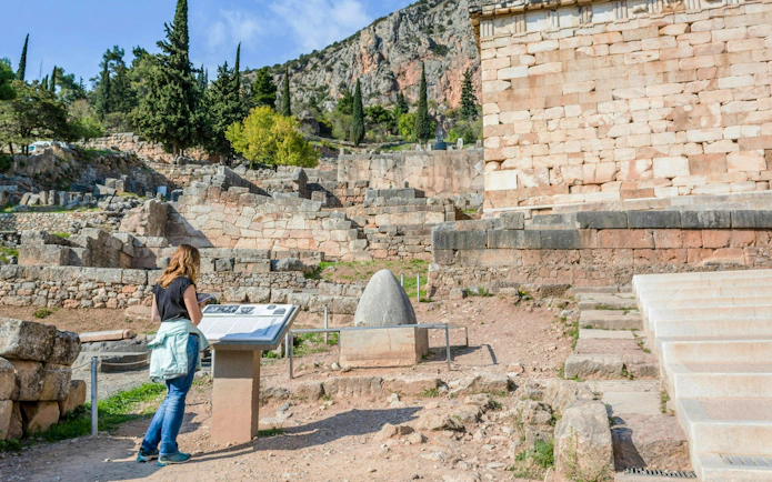 Visitor reading plaque at Delphi ruins, Greece, with ancient stone structures in background.