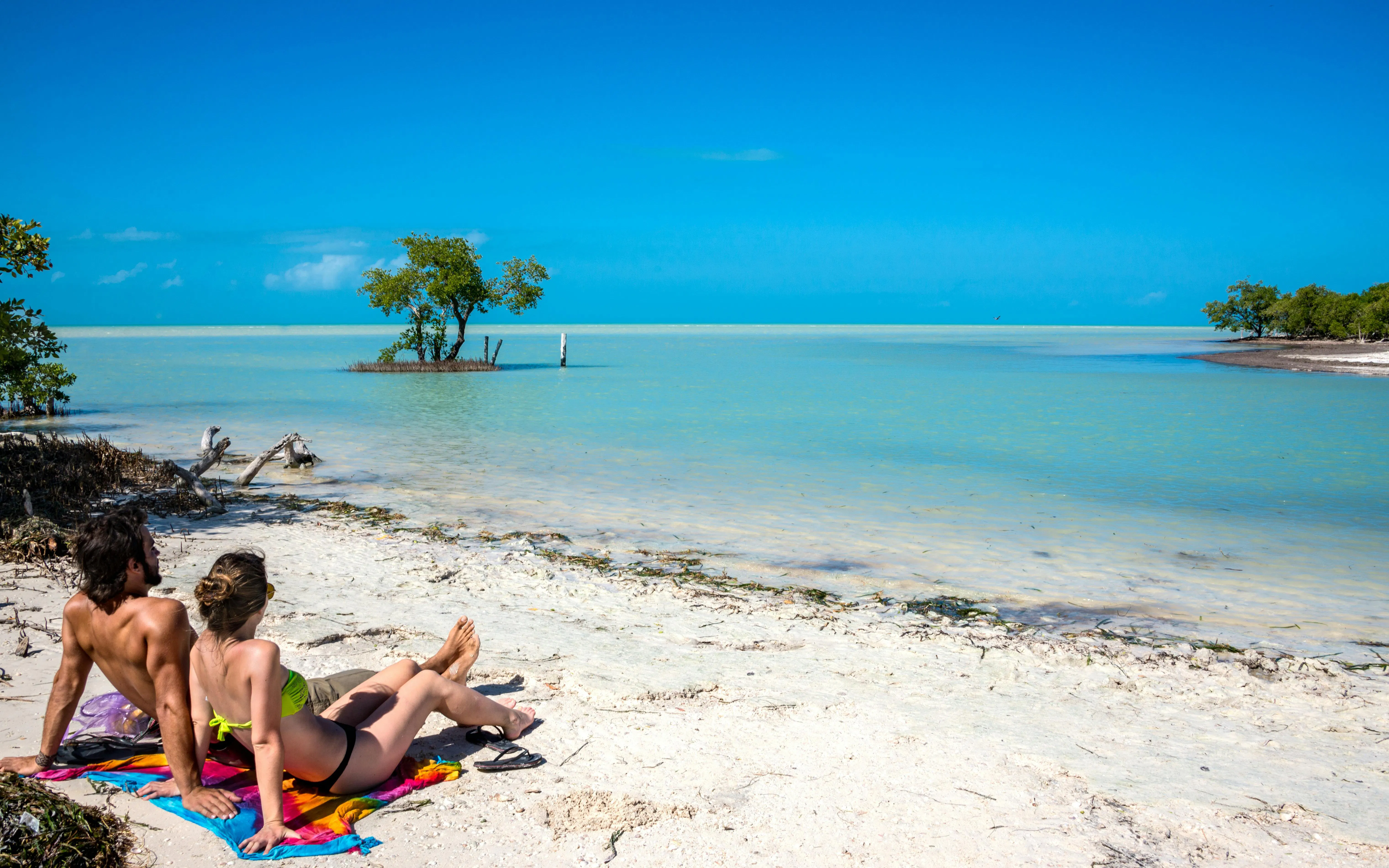 Couple relaxing on a sandy beach at Holbox Island, Mexico, with turquoise water and clear sky.