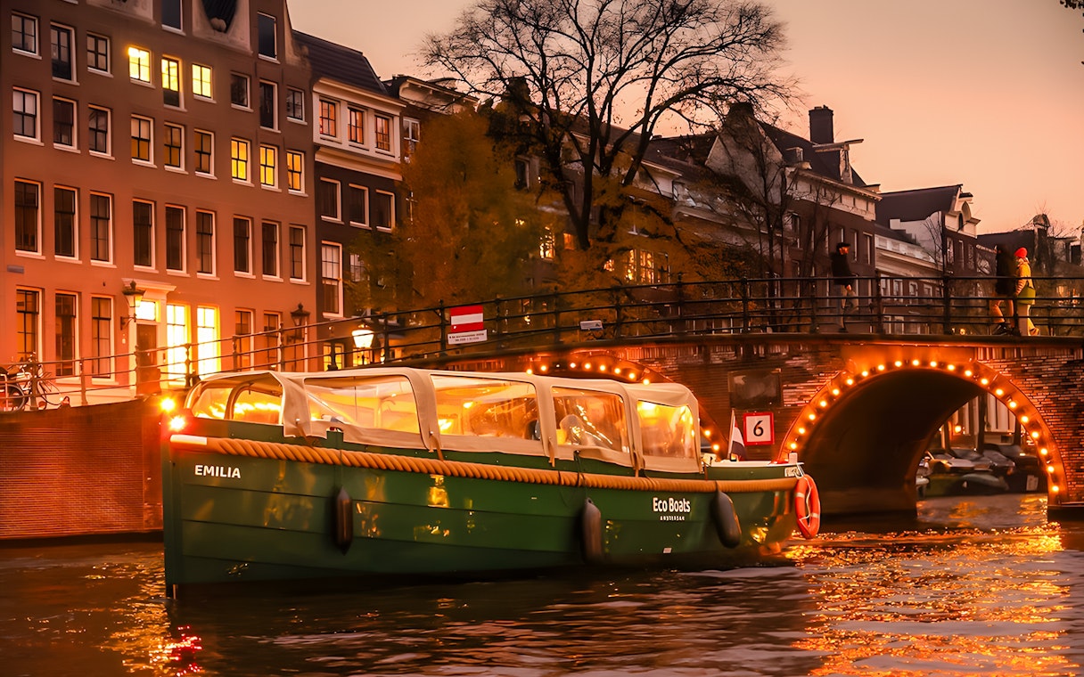 Amsterdam canal cruise boat with onboard bar passing under illuminated bridge at sunset.