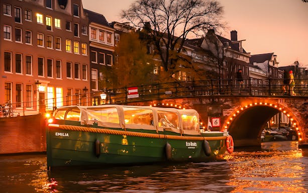 Amsterdam canal cruise boat with onboard bar passing under illuminated bridge at sunset.