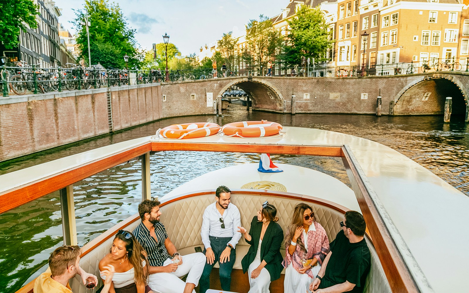 Open boat cruising through Amsterdam canals with passengers enjoying drinks.