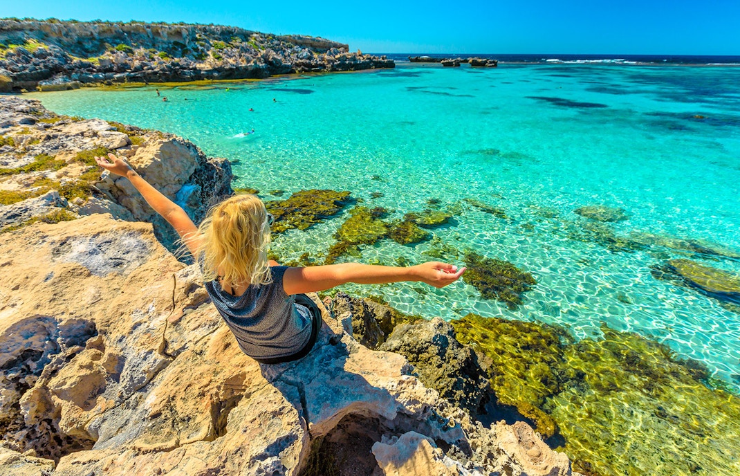 Person sitting on rocky shore overlooking clear turquoise water at a coastal lagoon.