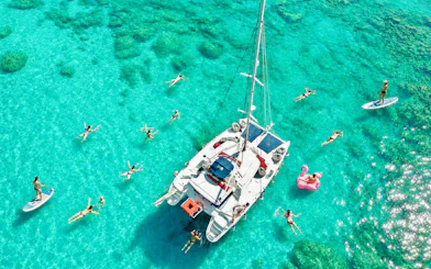 Catamaran surrounded by people swimming and paddleboarding in clear waters, Rhodes.