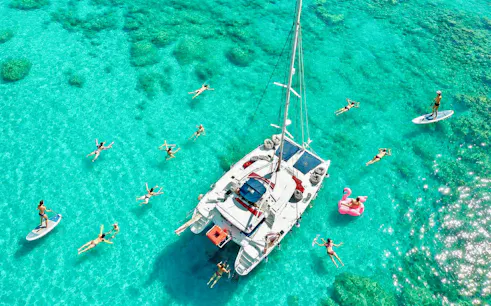 Catamaran surrounded by people swimming and paddleboarding in clear waters, Rhodes.