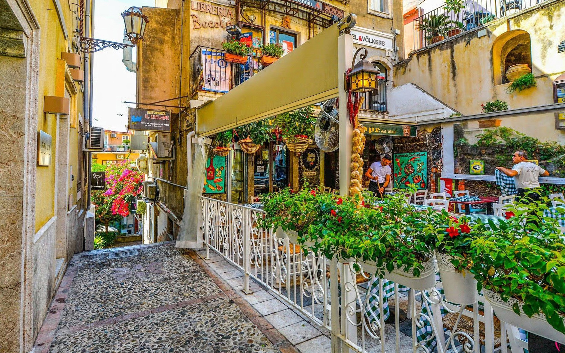 Narrow alleyway in Taormina with outdoor café and vibrant flowers.