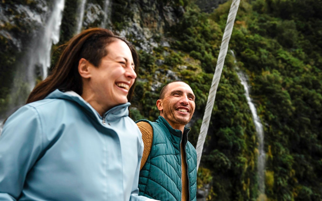 Couple laughing on a boat with waterfall and cliffs in Doubtful Sound, New Zealand.