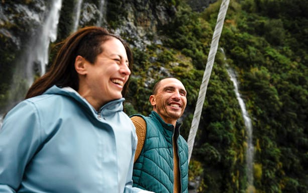 Couple laughing on a boat with waterfall and cliffs in Doubtful Sound, New Zealand.