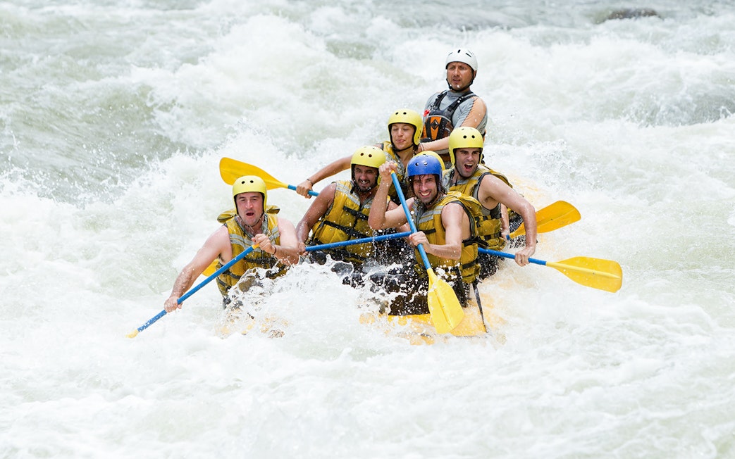 Group rafting through whitewater rapids with a guide, holding yellow paddles.