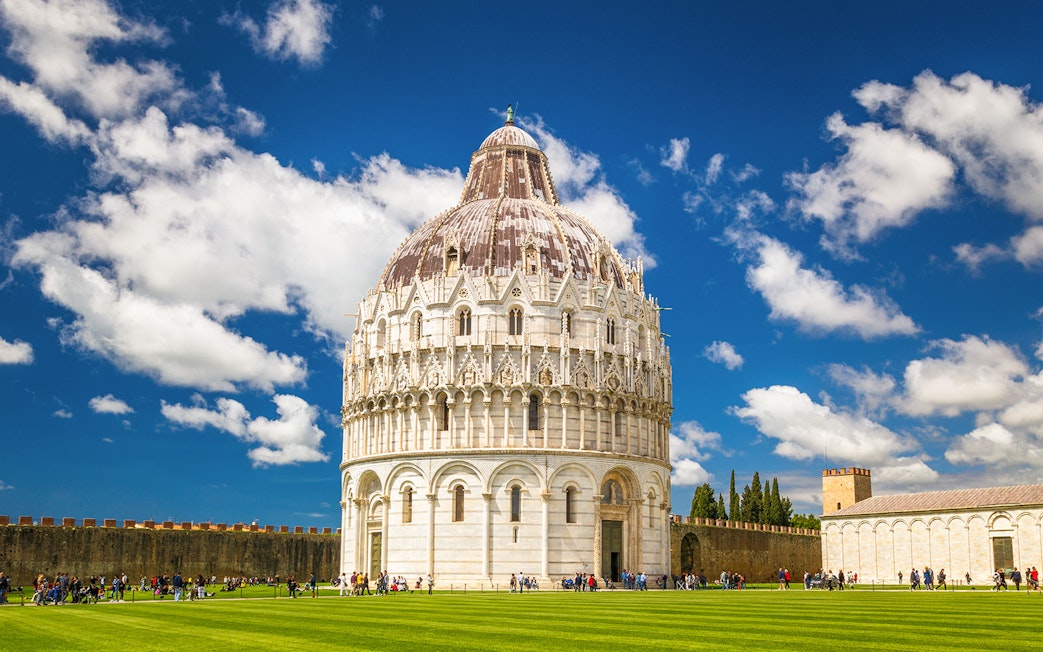 Pisa Baptistery with tourists on green lawn under blue sky.