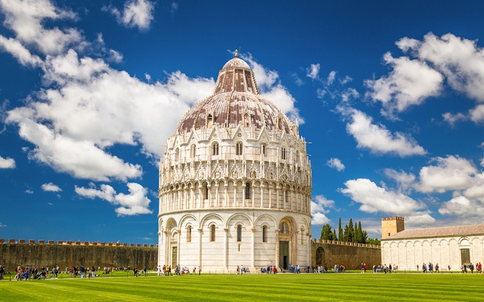 Pisa Baptistery with tourists on green lawn under blue sky.