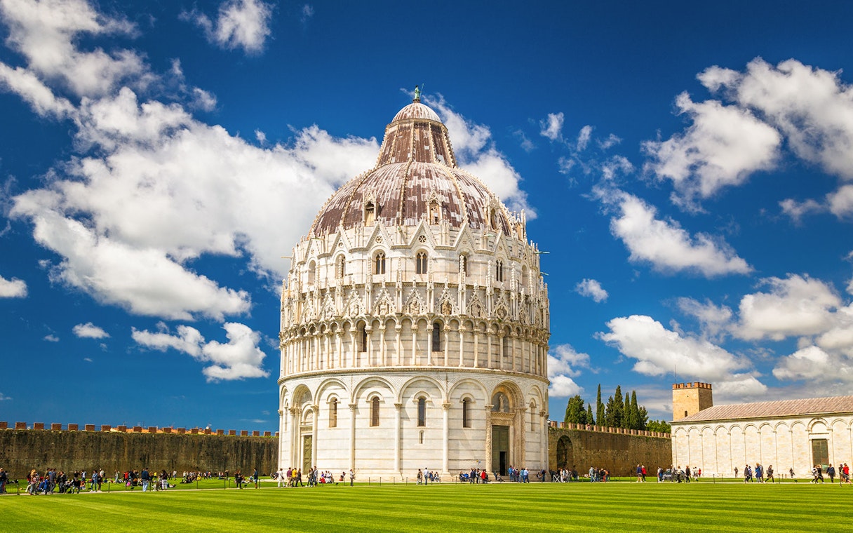 Pisa Baptistery with tourists on green lawn under blue sky.
