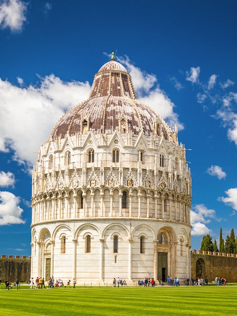 Pisa Baptistery with tourists on green lawn under blue sky.