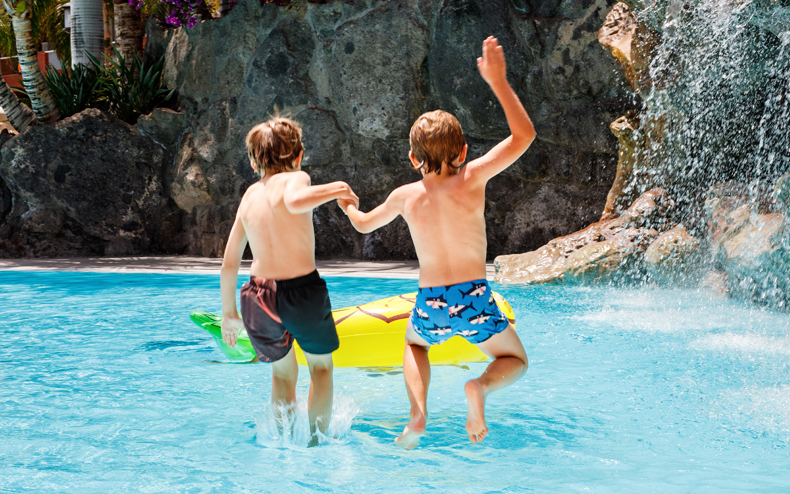 Kids jumping into pool at water park with waterfall and inflatable float.